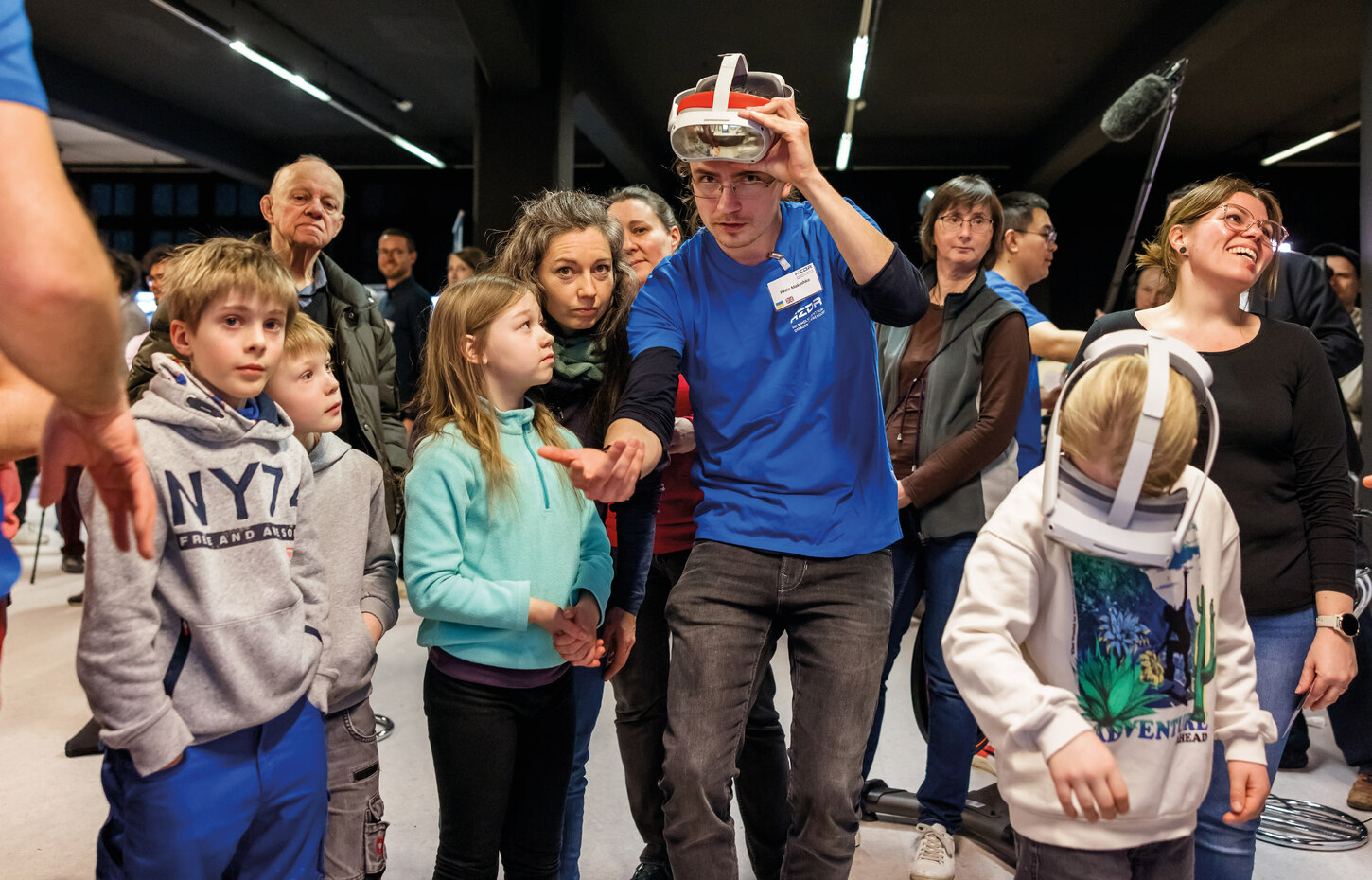 Einige Kinder und Erwachsene blicken interessiert einen Mann mit blauem T-shirt an, der eine technische Brille auf dem Kopf trägt und gestikuliert.